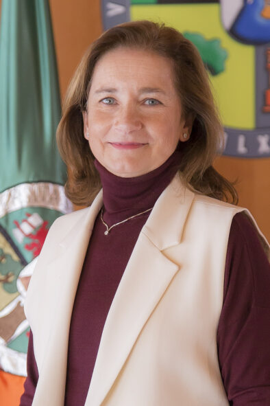 Foto oficial tipo retrato de Mª Mercedes Macías García delante de una pared con la bandera y el logo de la universidad.